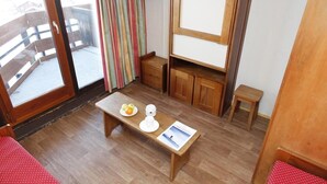 A compact room with a wooden floor, a small table with a plate of fruit, a white cup, and a notebook. There is a sliding glass door leading to an outdoor area with a wooden deck. - Studio Cosy + Accès Direct aux Pistes! (Saint-Martin-de-Belleville)