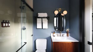 Un baño moderno con cabina de ducha de vidrio, inodoro blanco, tocador de madera con espejo y lavamanos blanco.