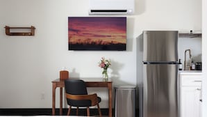 Habitación de hotel con cama, escritorio con silla, refrigerador, basurero, aire acondicionado de pared y un cuadro de un atardecer.