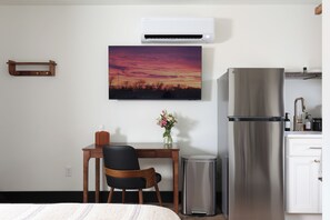 Et hotelværelse med en seng, et skrivebord med stol, et køleskab, en skraldespand, en vægmonteret aircondition og et billede af en solnedgang.