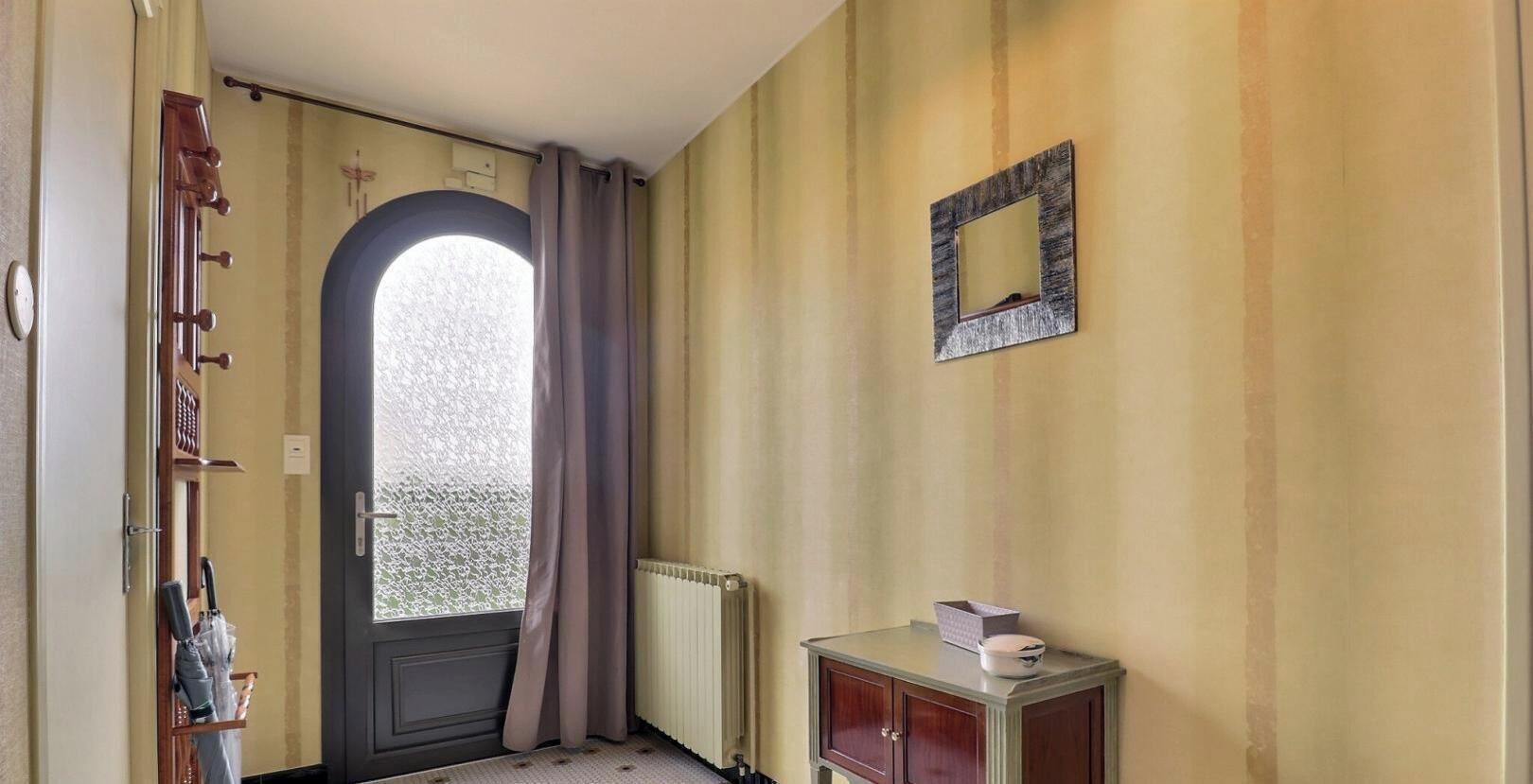 Ein Zimmer mit Tür, einem Fenster mit Vorhängen, einem Heizkörper und einem kleinen Schrank mit Gegenständen darauf.