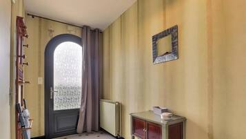 Ein Zimmer mit Tür, einem Fenster mit Vorhängen, einem Heizkörper und einem kleinen Schrank mit Gegenständen darauf.