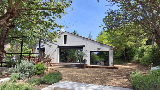 A modern house with a large glass door, surrounded by a garden with various plants and a concrete pathway leading to the entrance. - Luxury house in the heart of the Luberon (Apt)