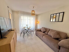 A living room with a TV, sofa, dining table, chairs, and a window with curtains. - Golfe Juan - 4 people - Swimming pool - Parking (Vallauris)