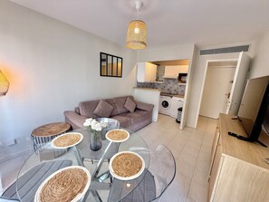 A modern living room with a glass dining table, a sofa, a TV, and a kitchen in the background. - Golfe Juan - 4 people - Swimming pool - Parking (Vallauris)