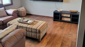 Un soggiorno con una grande TV a schermo piatto montata al muro, un divano in pelle marrone, un poggiapiedi a righe e un pavimento in legno.