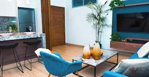 Una cocina moderna con encimera de mármol, dos taburetes de bar, una silla azul, una mesa de centro con bebidas, un televisor y una planta grande.