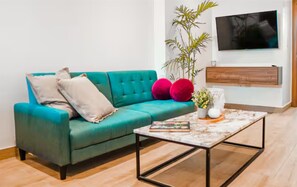 A teal sofa with cushions, a coffee table with books, a plant, and a TV on the wall.