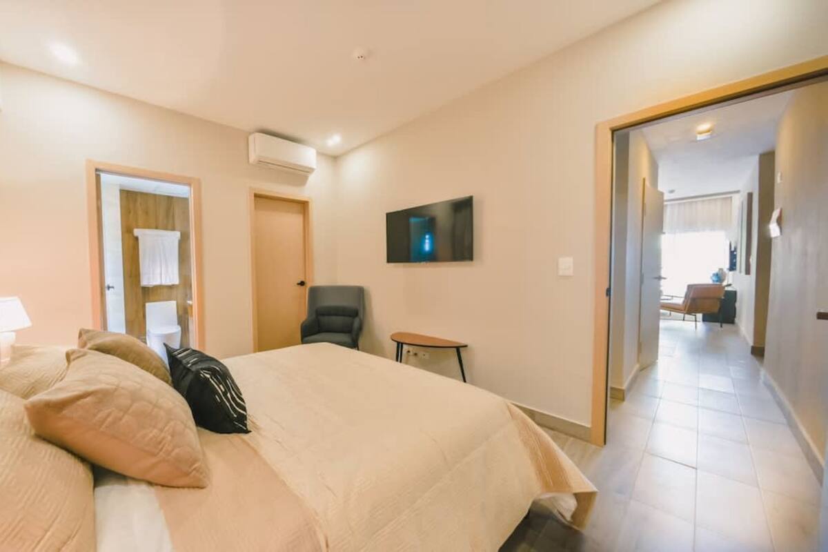 Ein Hotelzimmer mit Bett, Sessel, kleinem Tisch, Klimaanlage und Fernseher an der Wand.
