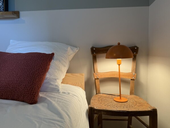 A bed with white bedding and a brown pillow, a wooden chair with a perforated seat, and a table lamp with an orange shade.