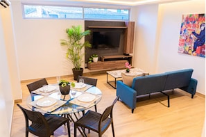 A dining area with a round glass table, chairs, a sofa, a TV, and a potted plant.