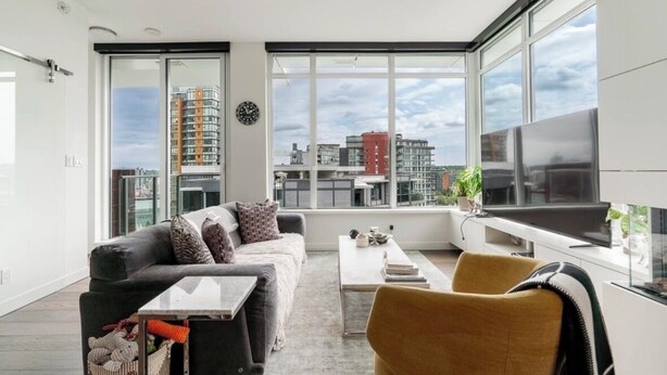 Una sala de estar moderna con sofá, mesa de centro y televisor. Amplias ventanas ofrecen vistas de la ciudad.