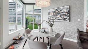 Un comedor moderno con una mesa redonda, sillas, una araña, un cuadro abstracto grande y vistas de edificios a través de amplios ventanales.