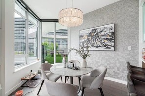 Un espace de salle à manger moderne avec une table ronde, des chaises, un lustre, une grande peinture abstraite et une vue sur les bâtiments à travers de grandes fenêtres.