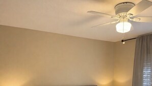 A hotel room with a bed, two bedside lamps, a ceiling fan, a small stool, and a window with curtains. - Charming 3-bedroom house in the heart of Palm Desert! (Palm Desert)