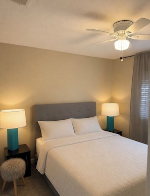 Habitación de hotel con una cama, dos lámparas de noche, un ventilador de techo, un taburete pequeño y una ventana con cortinas.