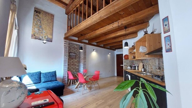 A modern interior with exposed wooden beams, a brick fireplace, a dining area with red chairs, and a kitchen with stone backsplash. - Nautilus - Cozy Apartment with Terrace in the City Center (Cagliari)