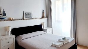 A bedroom with a wooden ceiling, a bed with a black headboard, a bedside table, a nightstand, a window with curtains, and a wall-mounted light fixture. - Nautilus - Cozy Apartment with Terrace in the City Center (Cagliari)