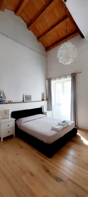 Une chambre avec un plafond en bois, un lit doté d’une tête de lit noire, une table de chevet, une commode, une fenêtre avec des rideaux et un luminaire fixé au mur.