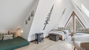 Ein gemütliches Schlafzimmer mit schrägen Wänden, einem Bett mit grüner Decke, einem Wandheizkörper und einem Fenster mit Blick auf Bäume.