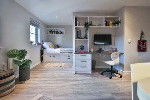 A modern bedroom with a bed, a desk, a chair, and a TV. There are shelves with books and plants, and a window with blinds. - Student Only Percys Place (York)