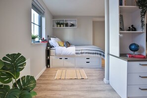 A bedroom with a bed, bedside tables, a window, a shelf with books and plants, and a large plant in the corner. - Student Only Percys Place (York)