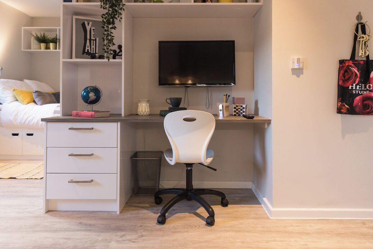 A bedroom with a bed, a desk with a chair, a TV, and a shelf with books and decorative items.
