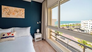 Ein Hotelzimmer mit Bett, Nachttisch, Lampe, Kopfkissen mit 'masterhost'-Stickerei, einem Bild und Blick auf Strand und Straße.