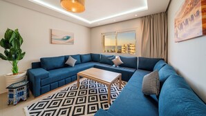 Ein modernes Wohnzimmer mit einem Ecksofa, einem Couchtisch, einer großen Kübelpflanze, einem gemusterten Teppich und Blick auf die Stadt durch ein Fenster.