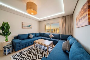 Ein modernes Wohnzimmer mit einem Ecksofa, einem Couchtisch, einer großen Kübelpflanze, einem gemusterten Teppich und Blick auf die Stadt durch ein Fenster.