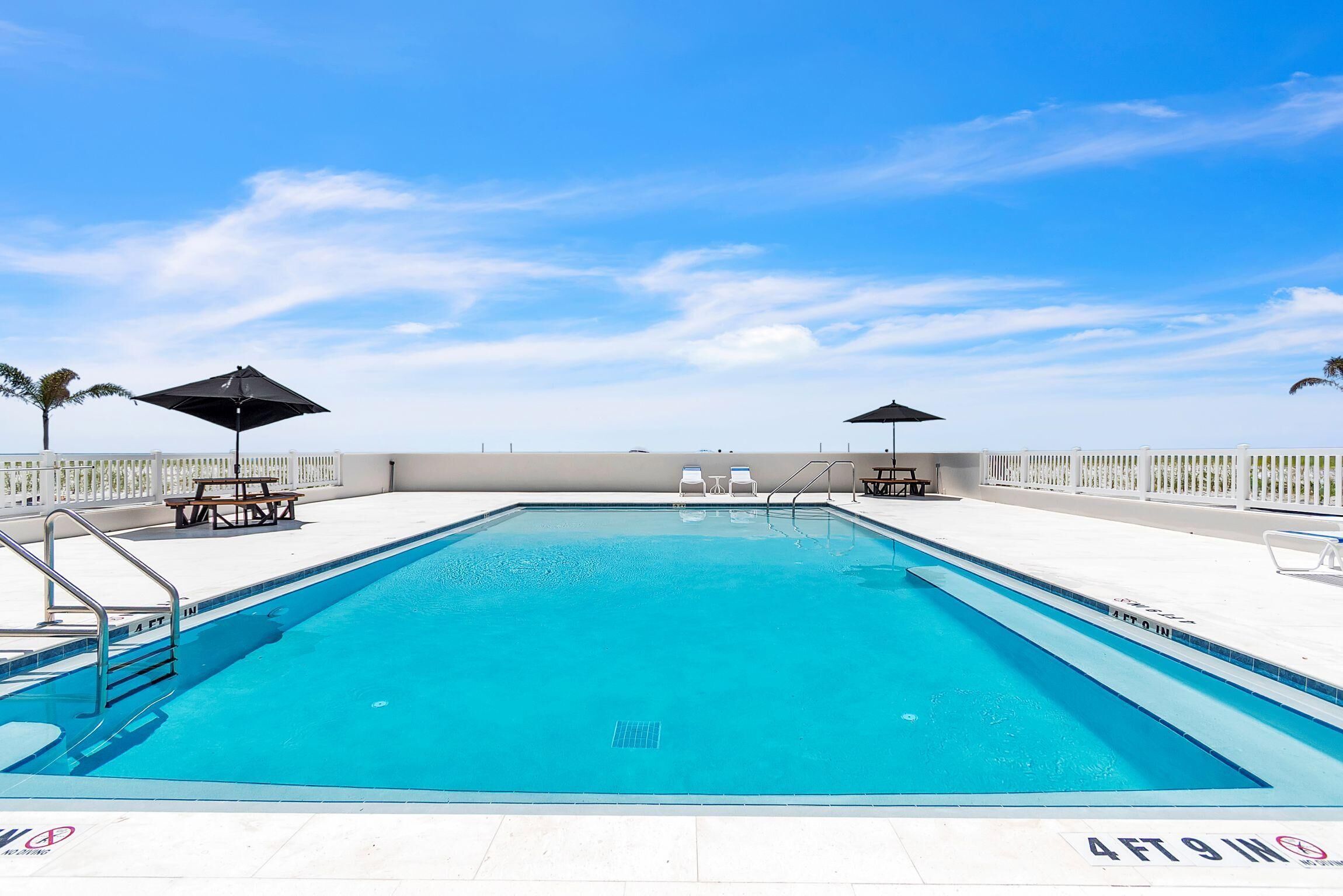A rectangular swimming pool with clear blue water, surrounded by a white deck with lounge chairs and umbrellas.