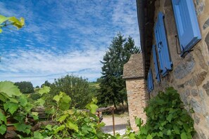 Ein Steinhaus mit blauen Fensterläden, umgeben von Grünpflanzen und Bäumen.