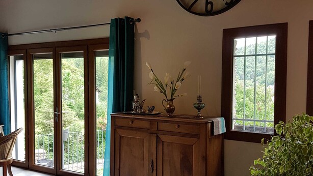 A room with a large wall clock, wooden cabinet, potted plant, and a view through a window. - GÎTE GABRIELLE - BAGNOLS-LES-BAINS (BAGNOLS-LES-BAINS)
