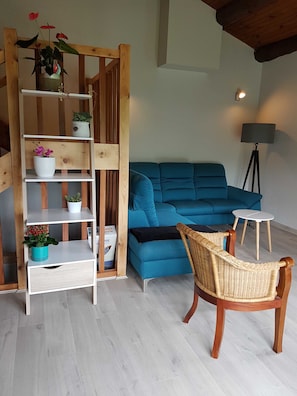 Un salón con un sofá azul, una silla de mimbre, una estantería de madera con plantas y una pequeña mesa blanca.