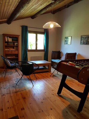 Una habitación con una mesa de madera, sillas, una estantería, una mesa de fútbol y una ventana con cortinas, además de una lámpara colgante.