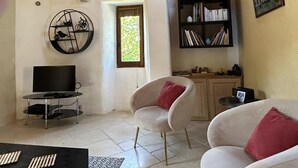 A living room with a TV, two white chairs with red cushions, a bookshelf, and a circular wall decoration. - Gîte de Poussiels - SAINT ANDEOL DE CLERGUEMORT (SAINT ANDEOL DE CLERGUEMORT)