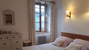 Ein Schlafzimmer mit Bett, Kommode, Fenster mit Vorhängen, Wandleuchte und Heizkörper.