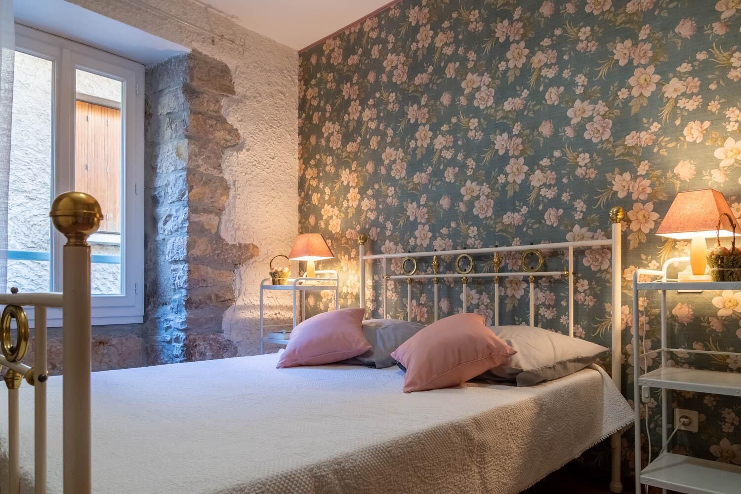 A bedroom with a stone wall, floral wallpaper, a bed with pink and white pillows, and two bedside lamps.