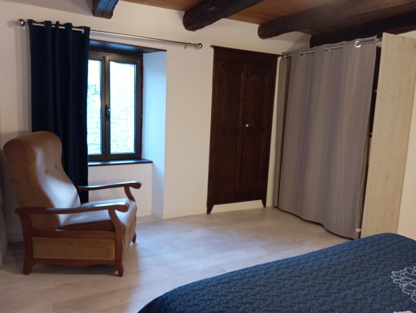 Een slaapkamer met een houten stoel, een raam met gordijnen, een kledingkast en een bed met een blauwe deken.