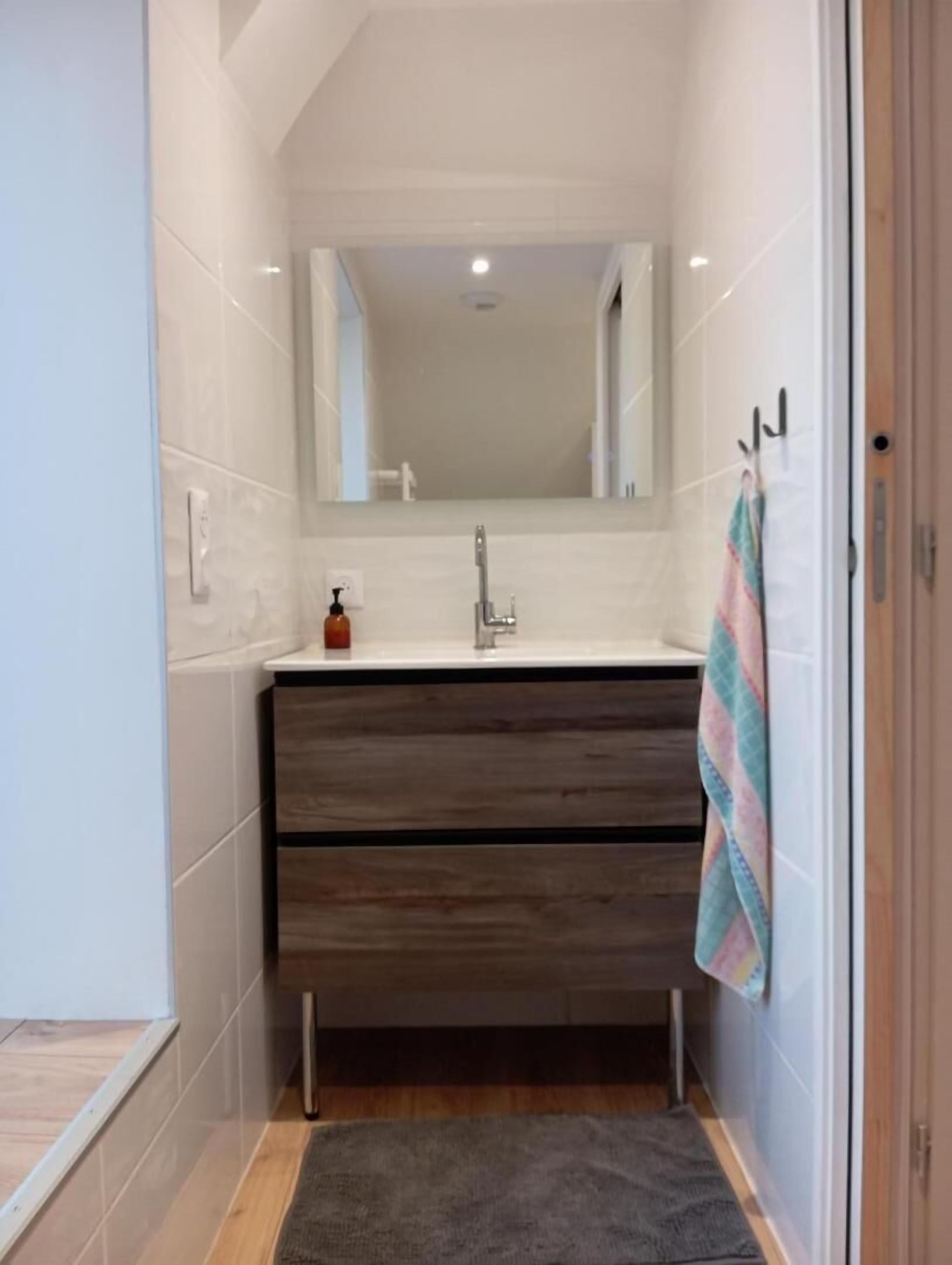 Ein schmales Badezimmer mit weiß gefliesten Wänden, einem hölzernen Waschtisch, einem Spiegel, einem Waschbecken und einem Handtuch, das an der Wand hängt.