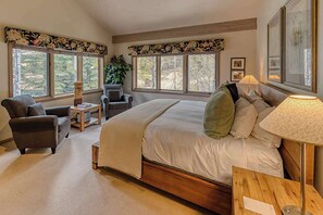 A bedroom with a large bed, two armchairs, a small table, a lamp, and a window with a view of trees.