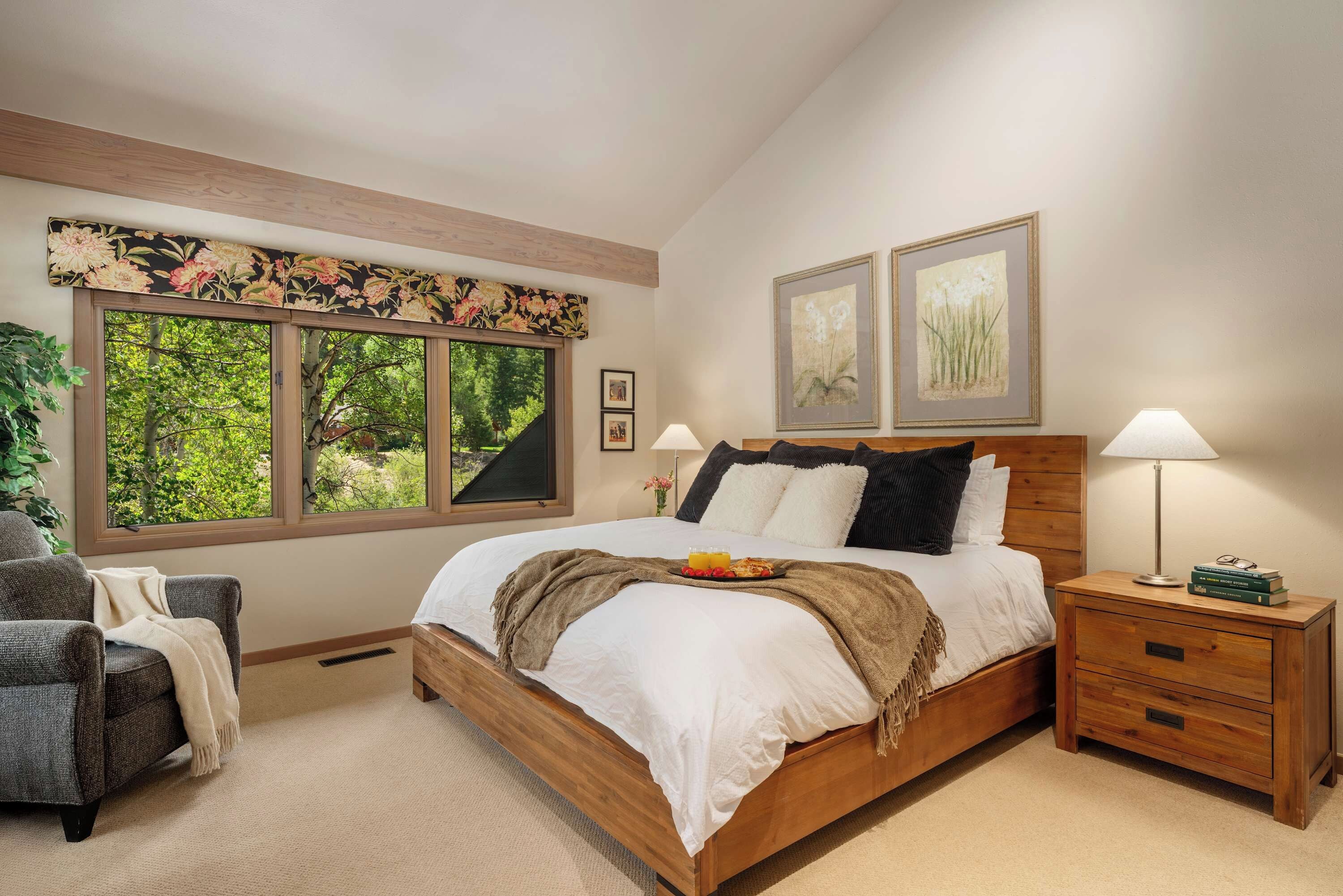 A bedroom with a large window, a bed with white linens, two bedside lamps, a nightstand, and a chair.