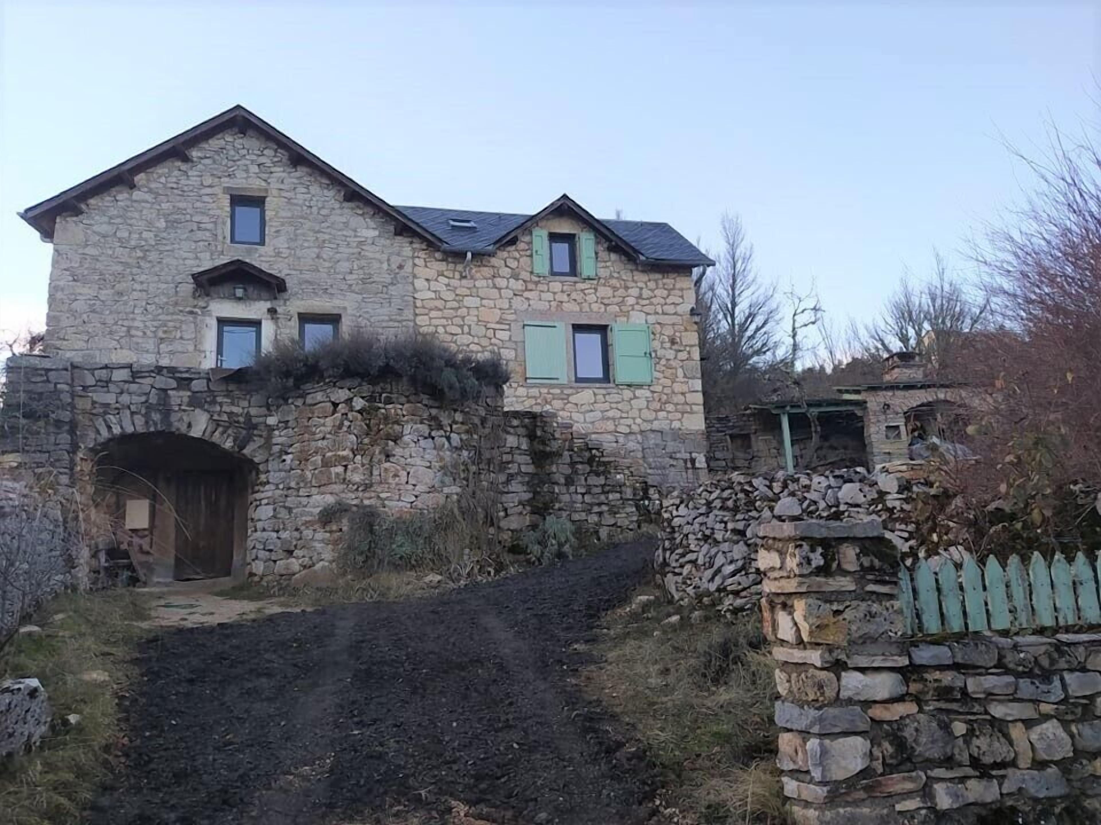 Una casa de piedra con techo inclinado, una puerta de madera y un muro de piedra con una reja.