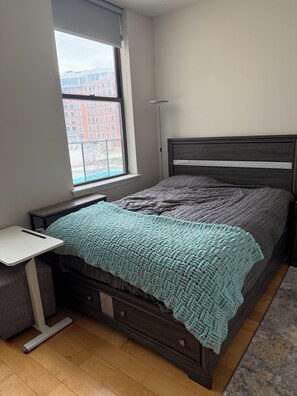 Ein Schlafzimmer mit einem Bett, einem Nachttisch, einer Stehlampe, einem Fenster mit Blick auf Gebäude und einem Holzboden.