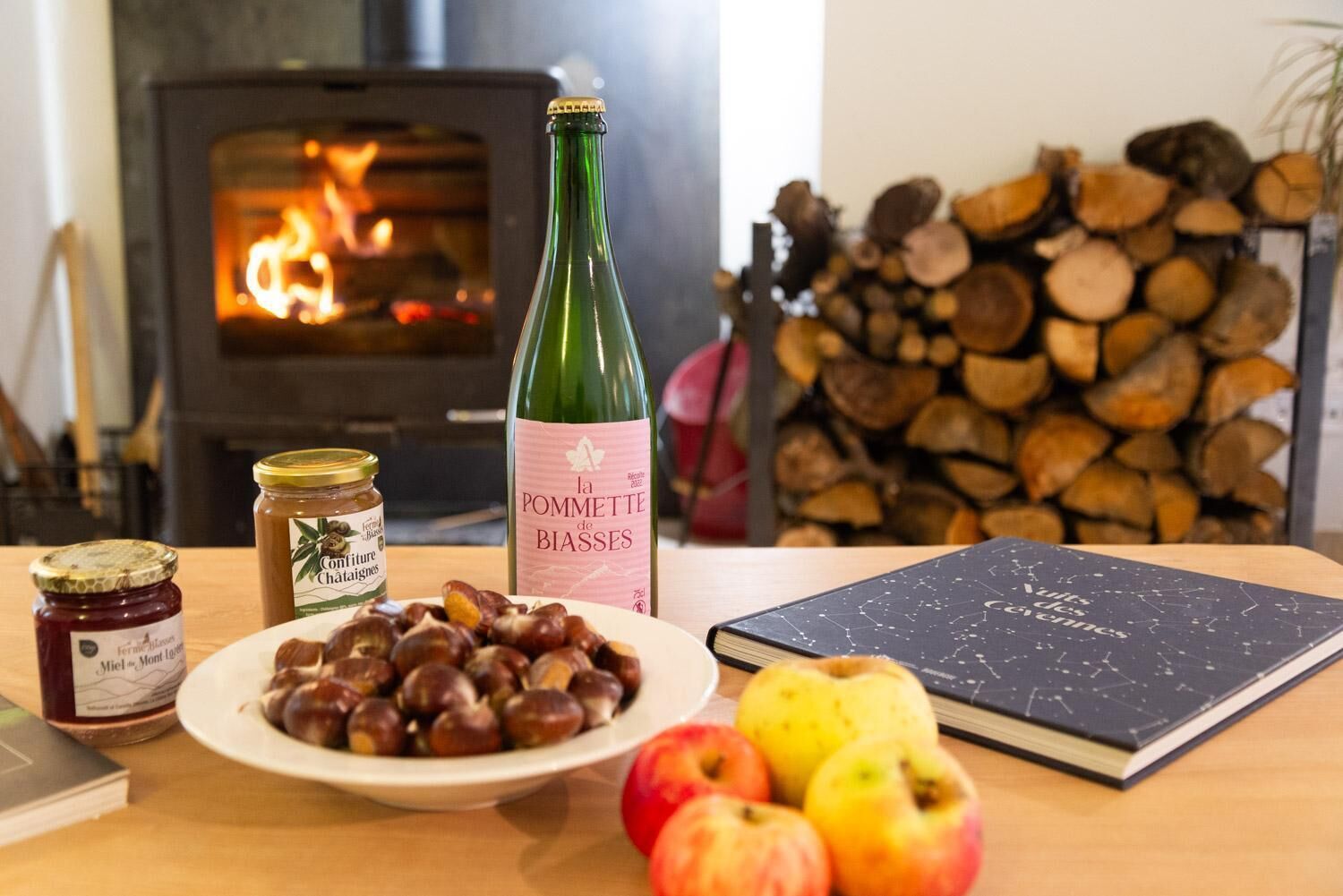 A table with a bottle of wine, a plate of chestnuts, apples, a book, and a jar of jam.