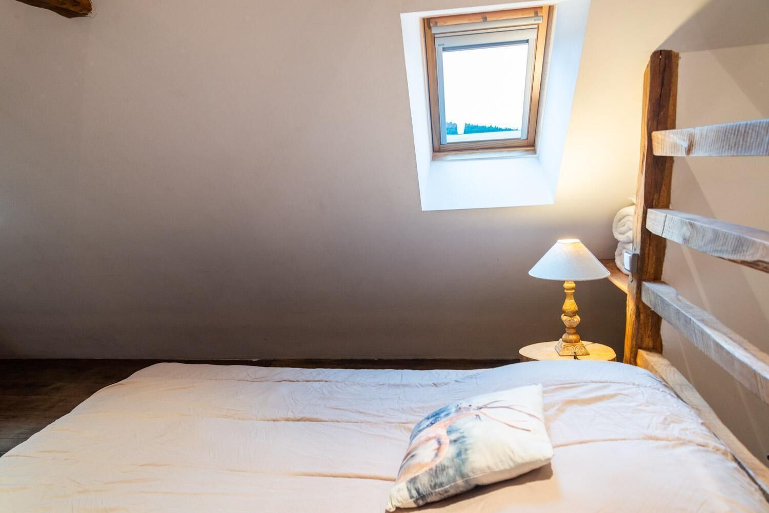 A bedroom with a single bed, a small table with a lamp, a window with a view, and a wooden headboard.