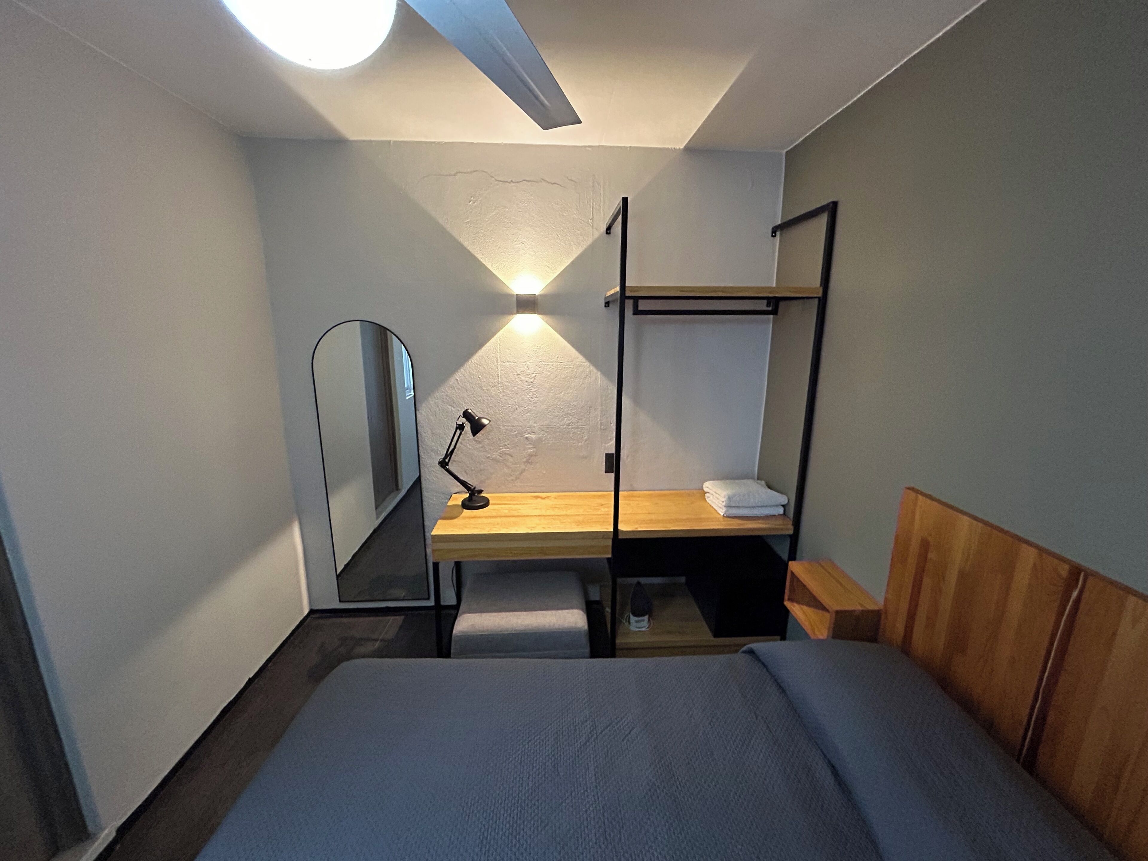 A compact hotel room with a bunk bed, a desk with a lamp, a mirror, and a bedside table.
