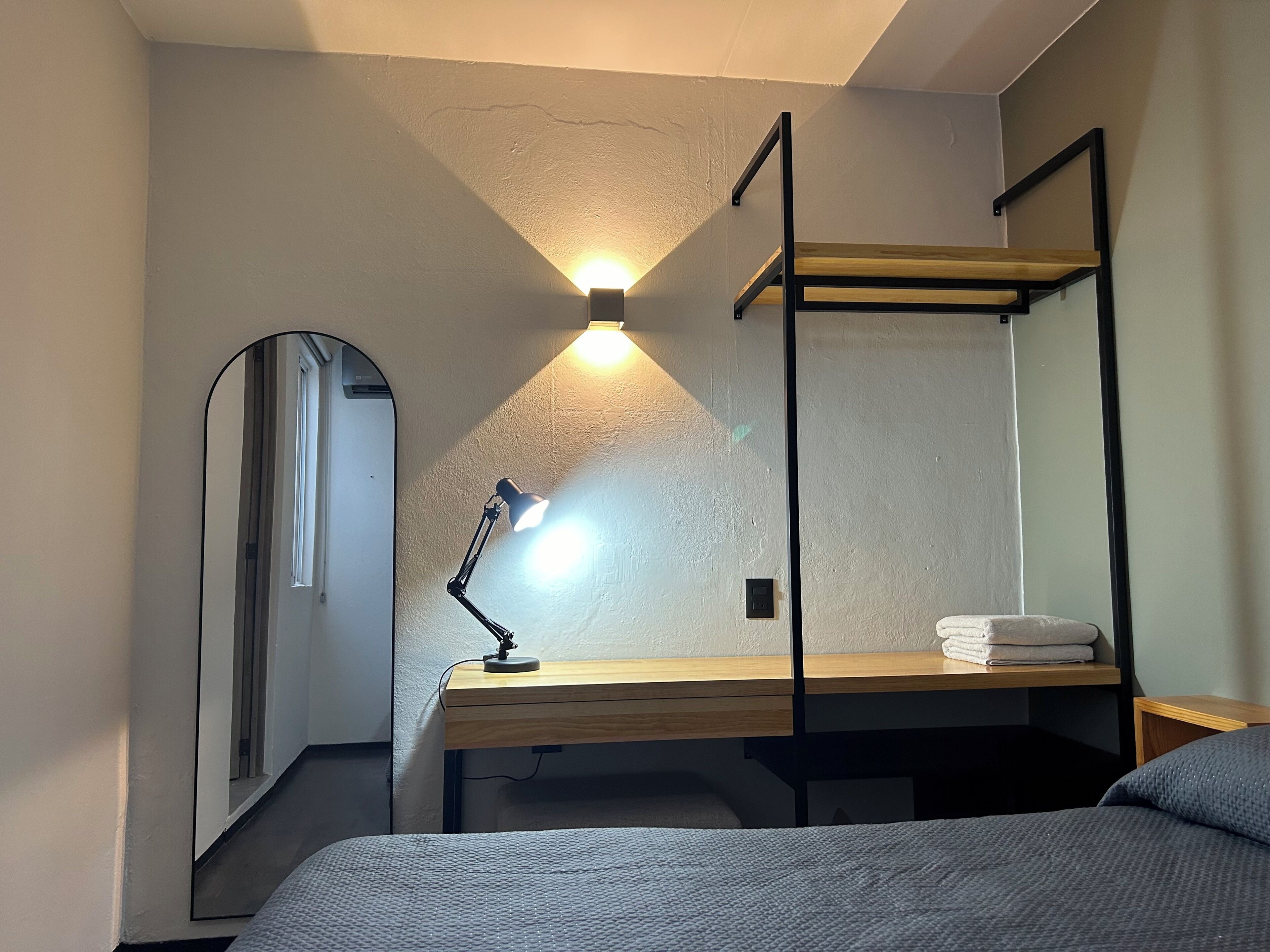 A compact room with a bunk bed, a desk with a lamp, a mirror, and a small shelf.