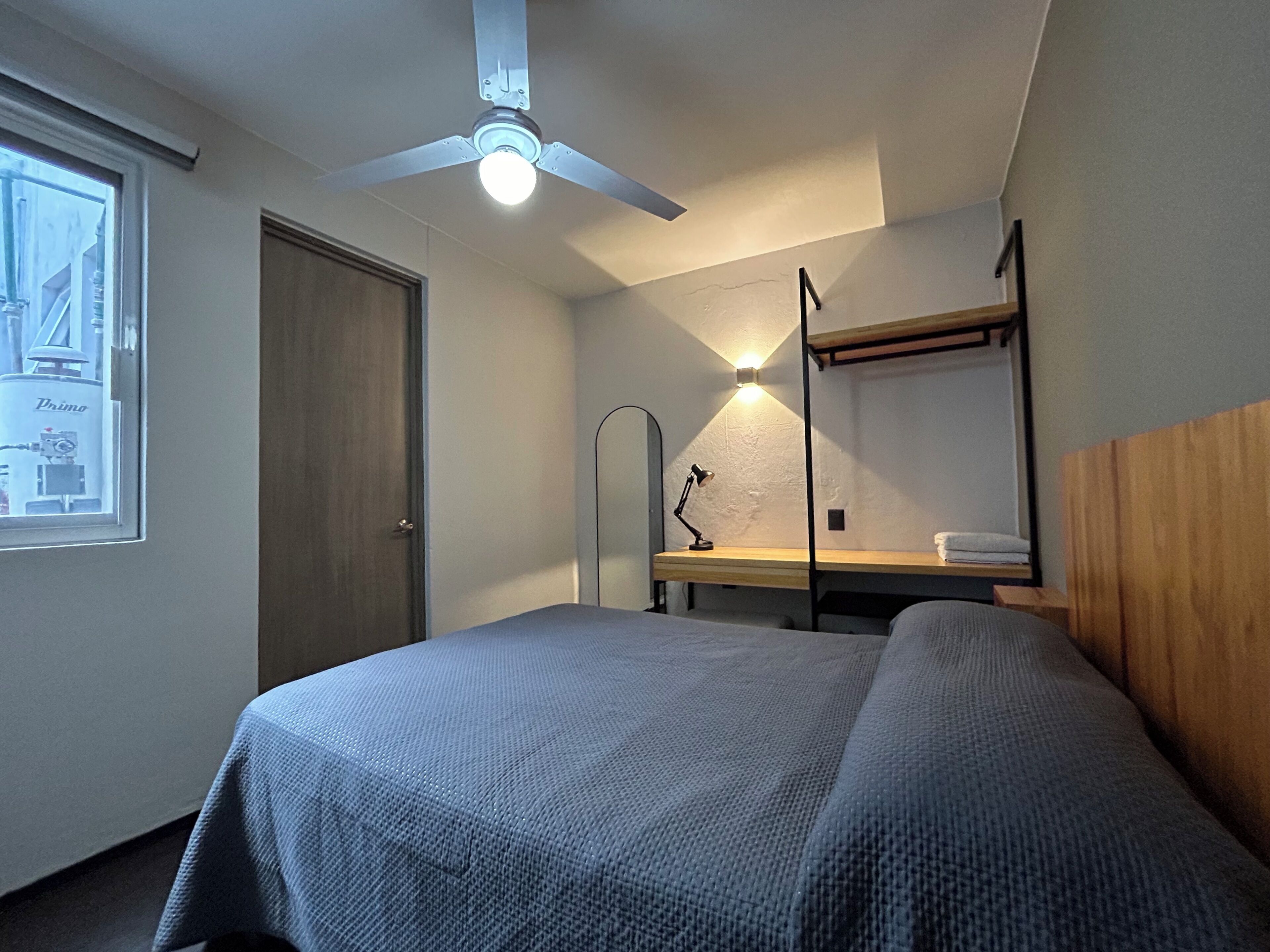 Um quarto com cama, escrivaninha, ventilador de teto, espelho e janela.