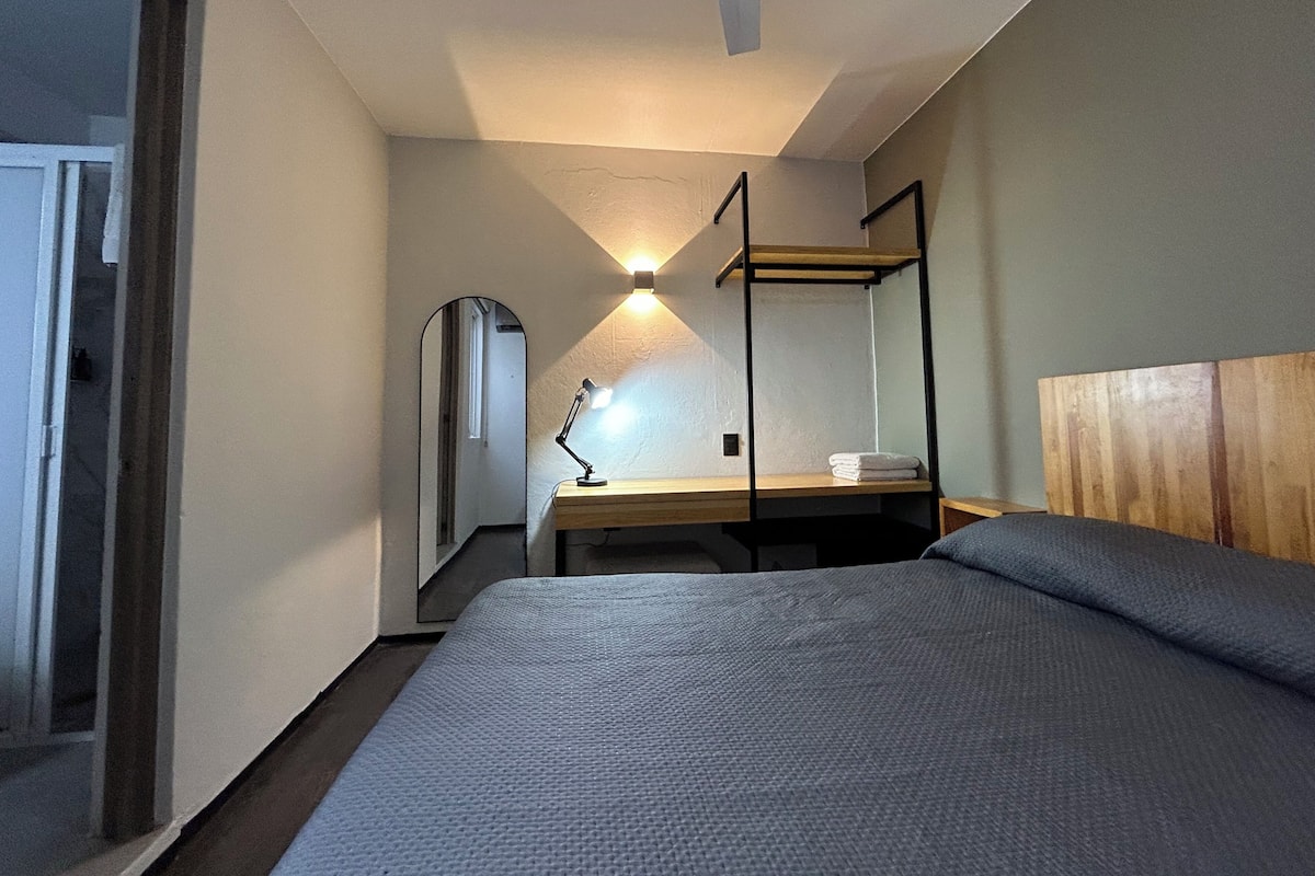 A bedroom with a bed, a desk with a lamp, a mirror, and a wooden headboard.